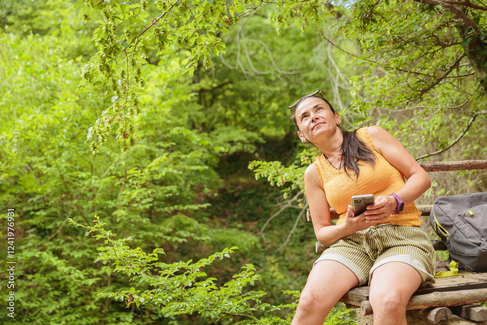 Woman using smartphone for pathfinding while relaxing sitting on wooden bench in forest Woman using smartphone for pathfinding while relaxing sitting on wooden bench in forest