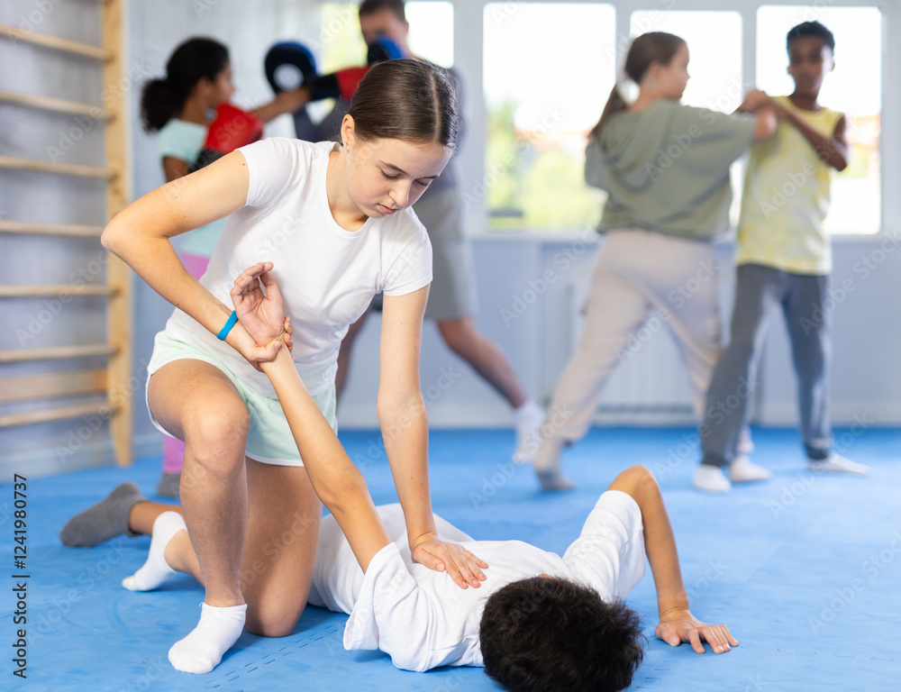 Focused teenage girl learning effective self defence techniques in sparring with boy, practicing painful armlock on opponent lying face down on floor in gym Focused teenage girl learning effective self defence techniques in sparring with boy, practicing painful armlock on opponent lying face down on floor in gym