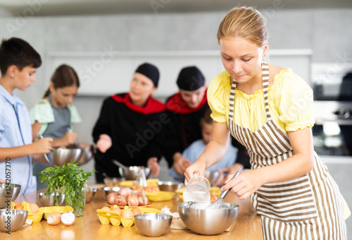 Interested enthusiastic teenage girl participating in culinary class of children group, engrossed in cooking process mirroring professional chef instructor steps photo