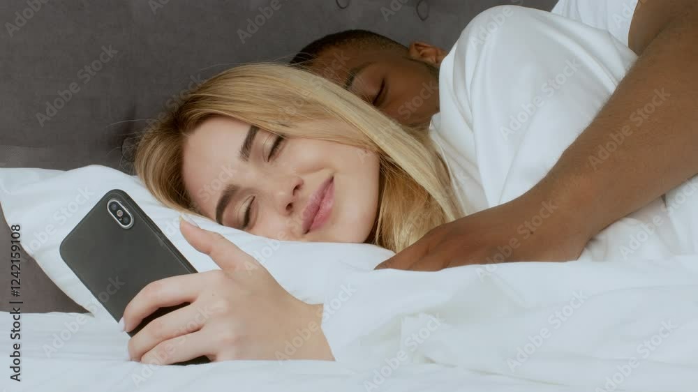 A young multiracial couple cuddles in bed, sharing a tender moment. The woman looks at her phone with a smile while the man embraces her from behind, creating a warm atmosphere. A young multiracial couple cuddles in bed, sharing a tender moment. The woman looks at her phone with a smile while the man embraces her from behind, creating a warm atmosphere.