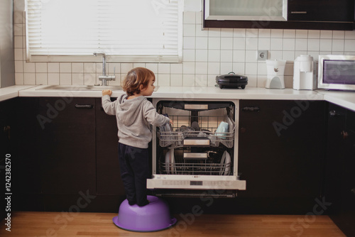 Child helps with dishes in modern kitchen while standing on stool photo