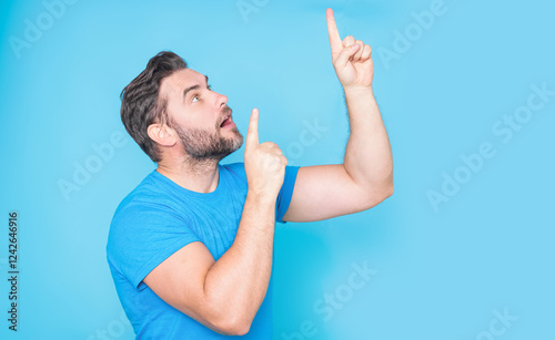 The point up. Look up. Happy handsome man pointing up away, isolated blue background. Excited man pointing a great idea. Guy in blue t-shirt pointing up with his finger. photo