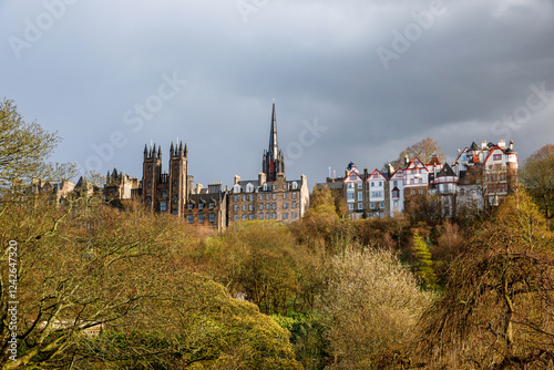 Obraz na płótnie Edinburgh skyline featuring historic architecture and gothic spires, framed by lush green trees under a dramatic cloudy sky. w salonie