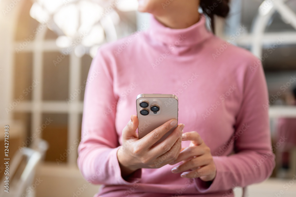 Fototapeta premium A thoughtful Asian woman gazes out the window while holding a smartphone, relaxing in a coffee shop.