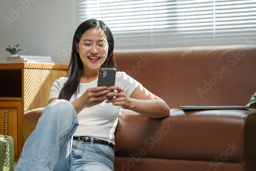 Smiling young woman is using her smartphone while sitting on a comfortable sofa in her living room, enjoying a relaxing moment at home with natural light streaming through the window
