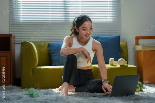 фотографія Smiling asian sportswoman sitting on carpet in living room using laptop searchin