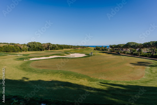 Golf course with a putting green, sand bunkers, trees, and ocean in the background under a bright blue sky, creating a serene view. photo