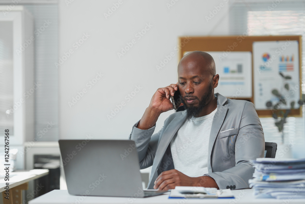 Serious african american businessman wearing gray jacket talking on cell phone and using laptop while analyzing documents at workplace in modern office Serious african american businessman wearing gray jacket talking on cell phone and using laptop while analyzing documents at workplace in modern office