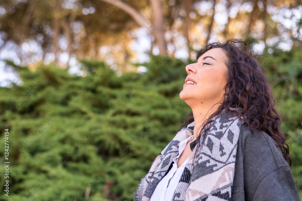 Serene woman with long, dark curly hair, wearing a shawl and a jacket, takes a deep breath of fresh air, embracing the tranquility and freedom of nature in a wooded park Serene woman with long, dark curly hair, wearing a shawl and a jacket, takes a deep breath of fresh air, embracing the tranquility and freedom of nature in a wooded park