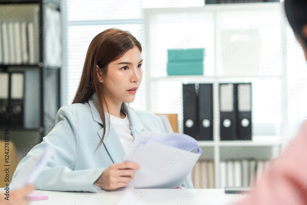 A focused woman sits at a table, holding documents, engaged in a discussion within a modern office setting filled with file organizers. A focused woman sits at a table, holding documents, engaged in a discussion within a modern office setting filled with file organizers.