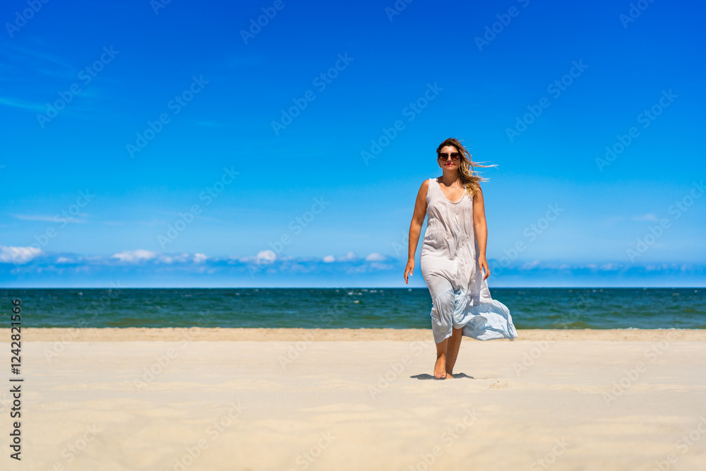 Beautiful middle aged woman in dress fluttering on wind walking on white sandy beach on sunny summer day. Front view Beautiful middle aged woman in dress fluttering on wind walking on white sandy beach on sunny summer day. Front view