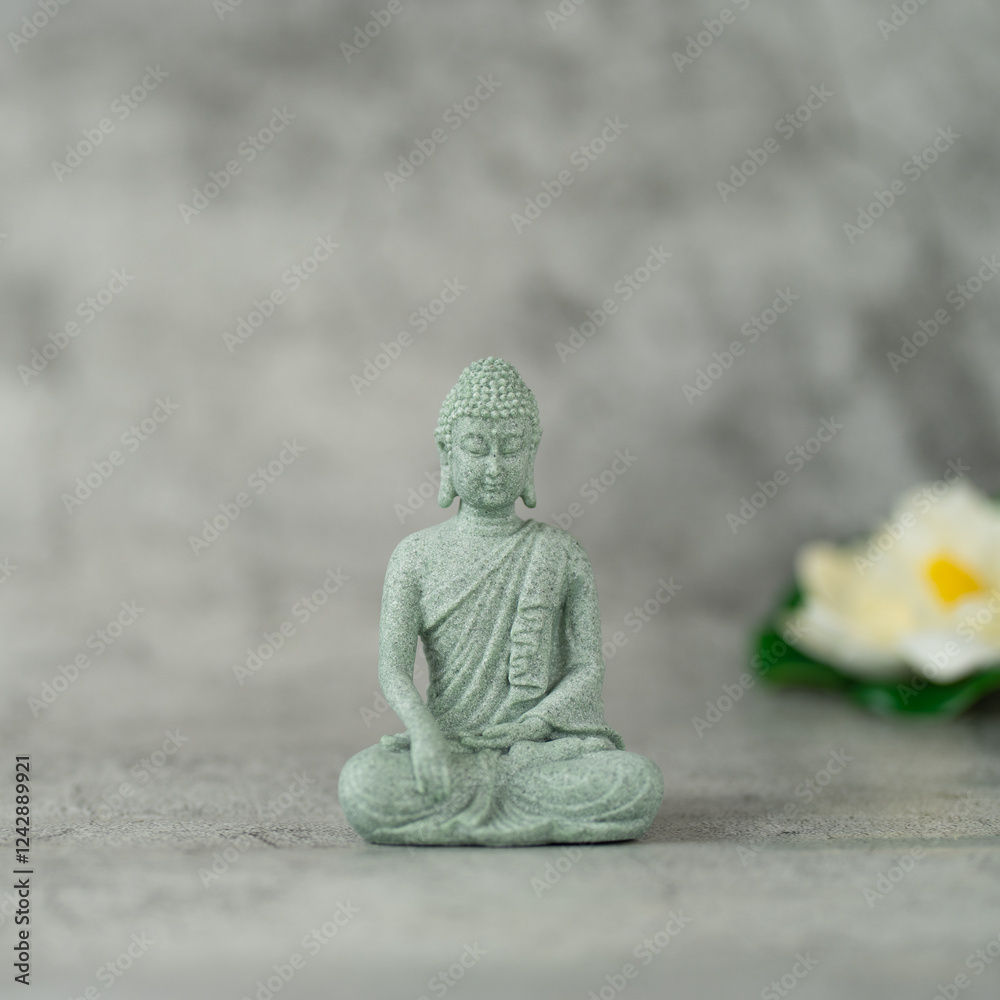 A small Buddha statue is sitting on a gray surface. The statue is made of stone and has a serene expression on its face. in the background an out-of-focus lotus flower is visible. A small Buddha statue is sitting on a gray surface. The statue is made of stone and has a serene expression on its face. in the background an out-of-focus lotus flower is visible.