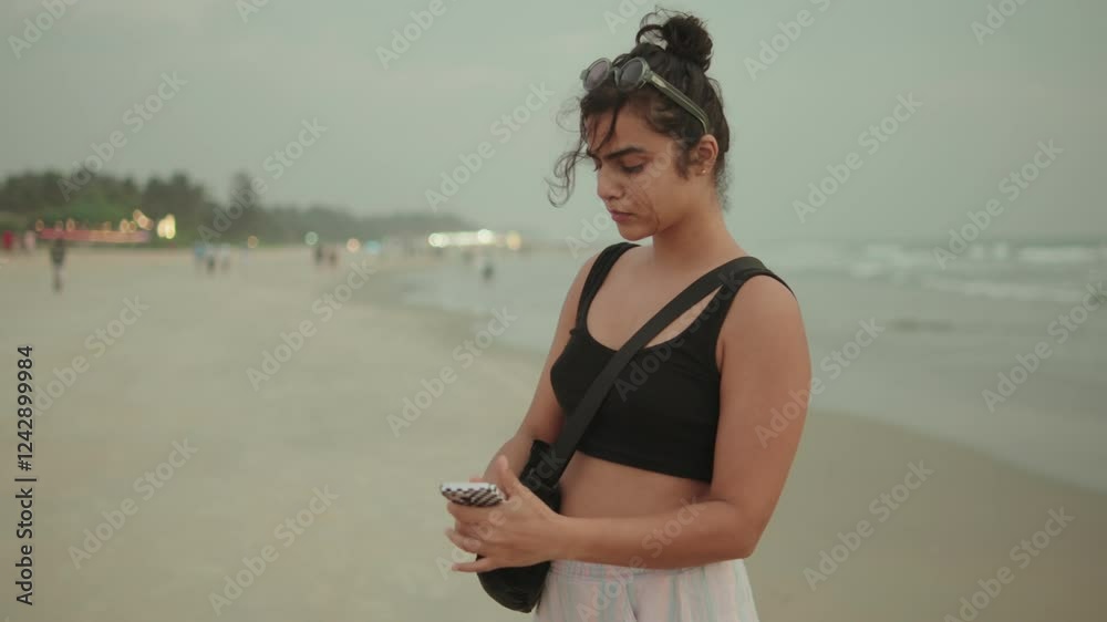 Young woman standing on the beach, holding a phone and looking out at the ocean Young woman standing on the beach, holding a phone and looking out at the ocean