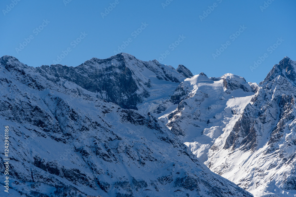 Obraz premium Dombay. Karachay-Cherkessia. Giant rocks with snow-capped peaks and glaciers rise majestically against blue autumn sky. Pine and spruce forests grow at foot of mountains. Sunny day. Autumn 2024