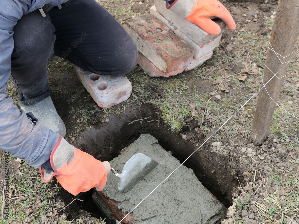 Obraz premium worker in orange gloves laying and leveling a layer of fresh cement with a trowel in a hole in the ground on a block with a string stretched nearby as a level, preparing for foundation work
