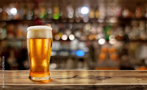 Fotografie Glass of chilled beer on wooden bar tabletop and blurred brightly lit bar interior in the background