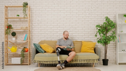 A man with a prosthetic leg reads a book sitting on a couch in a room with a modern home interior photo