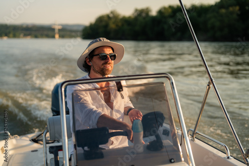 Young man enjoying a leisurely sunset cruise on a river, skillfully navigating a motorboat while dressed in a stylish white shirt, sunglasses, and a fashionable hat