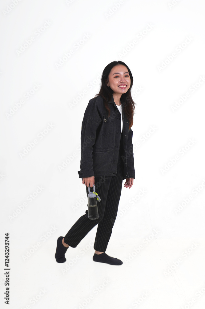 Standing or walking young woman with long hair wearing black denim jacket and pants holding reusable gray water bottle container isolated on vertical white background. Standing or walking young woman with long hair wearing black denim jacket and pants holding reusable gray water bottle container isolated on vertical white background.