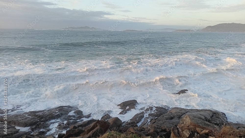 Obraz premium Stunning coastal scene featuring powerful ocean waves crashing against rugged rocks under a serene sky, with distant islands on the horizon. Perfect for nature, travel, and seascape themes