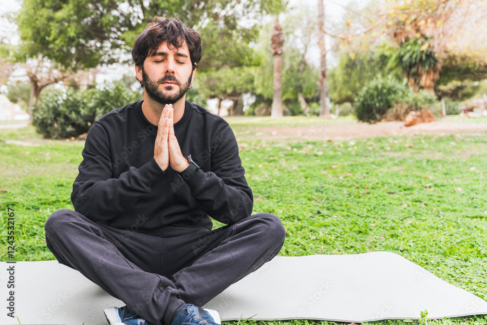 Peaceful bearded male practicing meditation, sitting cross-legged on yoga mat amid urban greenery