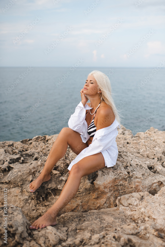 Attractive young adult woman 30-35 year old with long blonde hair wearing white shirt and swimsuit sitting on rocks over sea at background. Summer vacation season. Attractive young adult woman 30-35 year old with long blonde hair wearing white shirt and swimsuit sitting on rocks over sea at background. Summer vacation season.