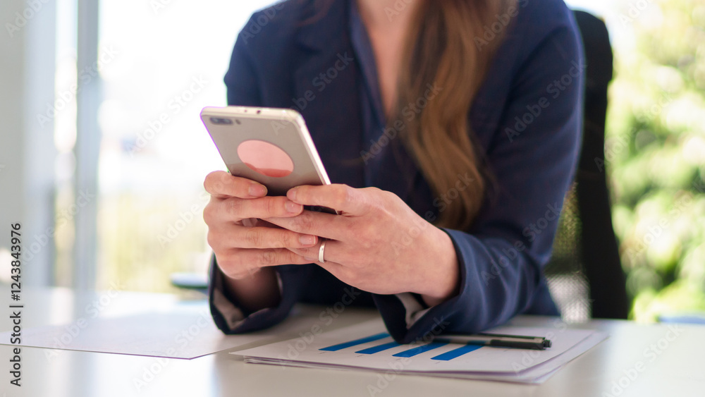 Fototapeta premium Close-up of a young Asian businesswoman using a smartphone while working in a bright and modern office