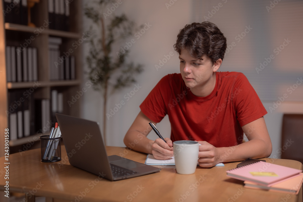 Young man attending an online class, sipping coffee and writing notes in a notebook while seated at a desk with a laptop, surrounded by books on a bookcase Young man attending an online class, sipping coffee and writing notes in a notebook while seated at a desk with a laptop, surrounded by books on a bookcase