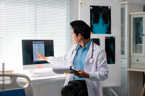 Doctor analyzing patient information form on computer screen, holding medical records in hand, in hospital room with x-rays on the wall