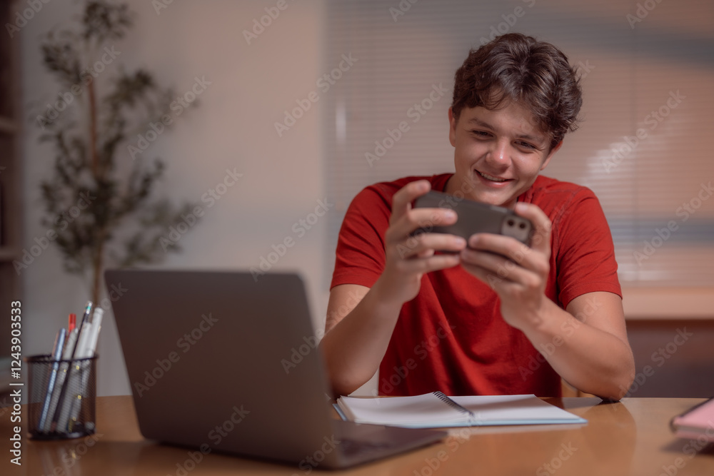 Young student is having fun playing mobile games on his phone instead of studying with his laptop and notebook, he is procrastinating and wasting time at his desk Young student is having fun playing mobile games on his phone instead of studying with his laptop and notebook, he is procrastinating and wasting time at his desk