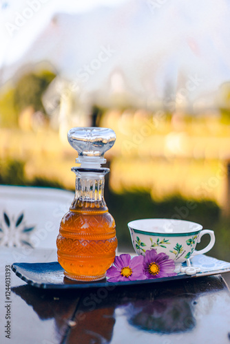 Close-up of a clear glass bottle with tea on a saucer. There is a floral patterned cup and a purple flower next to it. Blurred background of mountain views and farms with copy space. Feeling fresh.