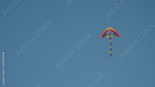 A rainbow kite with a long tail floats beautifully against a monochromatic blue sky.