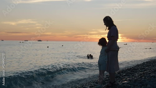 Mother and daughter standing on the beach on the seashore against the background of sunset. Mother and child embrace, after which the parent lifts the child in his arms.