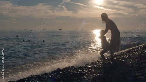 The mom is playing with her little girl on the sea beach. The girl is bouncing upwards over the sea waves coming at her, and the mother is helping the child to soar into the air.