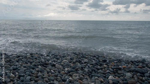 The sea waves crash one after another on the shore, made up of many pebbles of different colors and sizes. Above the sea is a gray sky with clouds.