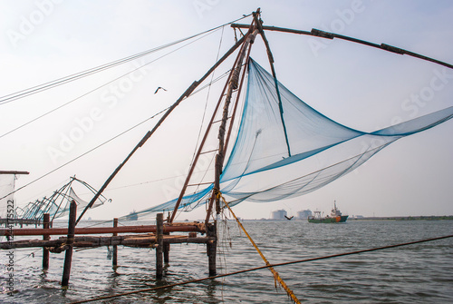 A traditional Chinese fishing net set in the water, stretching across the surface. A bird flies above while a cargo ship can be seen in the background, adding contrast to the rural fishing method.