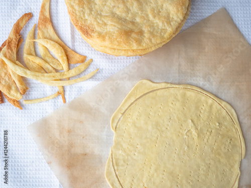 Cooking of a shortcrust puff pastry Napoleon cake with a pile of baked thin buttery layers, a raw layer rolled out on a baking paper and dough scraps on a white waffle towel.