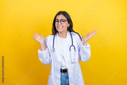 Young brunette doctor woman wearing stethoscope standing over isolated yellow background clueless and confused expression with arms and hands raised
