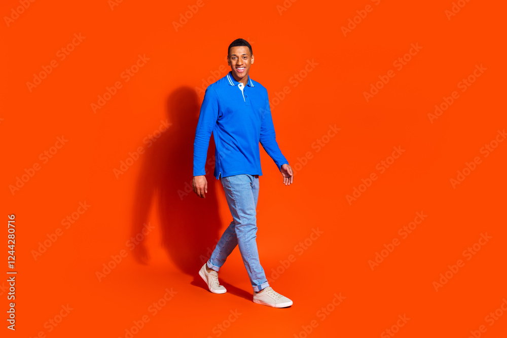 Smiling young man in blue walking confidently against bright orange background Smiling young man in blue walking confidently against bright orange background