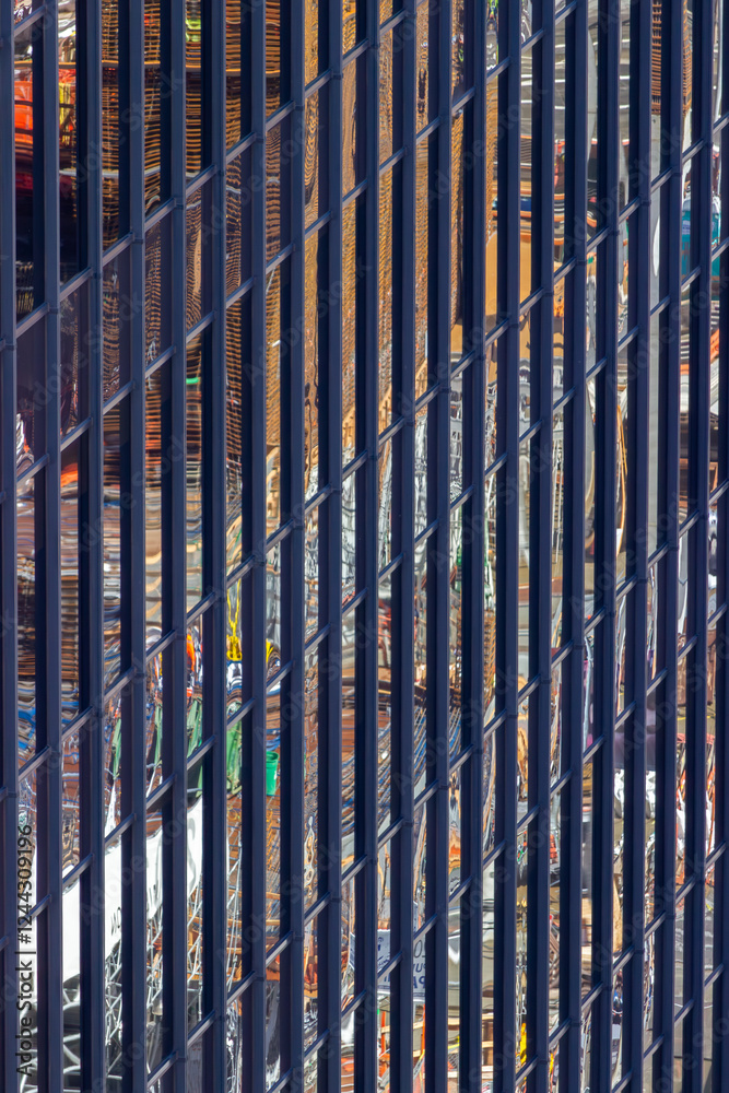 A close-up view of a modern building's facade, showcasing vertical metal bars with reflections of a vibrant cityscape, creating an abstract and dynamic visual effect.