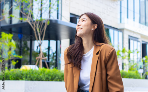 Carefree Young Asian Businesswoman looking up to sky, visualizing the future, relaxing, and dreaming about new journey in her life while sitting outdoors at the center of a city street building.