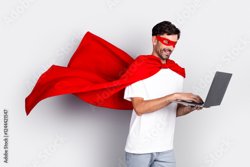 Cheerful man wearing a red superhero cape and mask using a laptop while standing against a white background