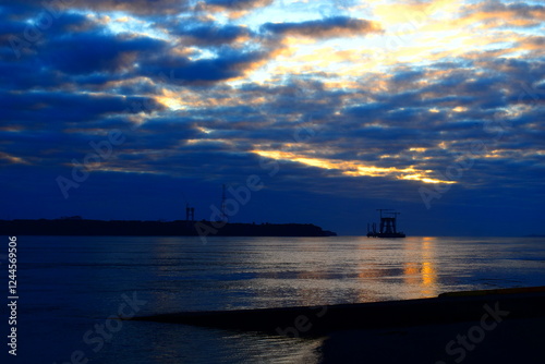 Breathtaking seascape at sunset with a dramatic sky filled with deep blue and golden clouds. The calm water reflects the warm light, creating a peaceful yet mysterious atmosphere. In the distance