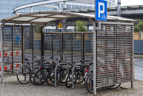 Bicycle parking shelter with multiple bikes locked to metal racks near a modern urban transport hub on a cloudy day. Eco-friendly transport, urban cycling and mobility