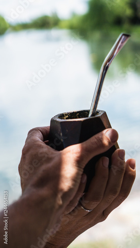 A hand holds a traditional mate cup with a metal straw, filled with yerba mate. The background features a blurred lake and lush greenery, creating a peaceful atmosphere.

