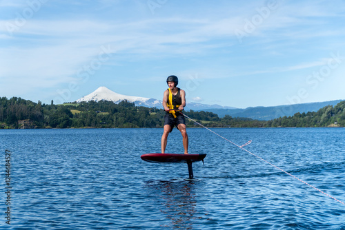 A young surfer wearing a helmet and life jacket, gliding on a foil board over a lake wave, towed by a boat with a majestic volcano in the background.