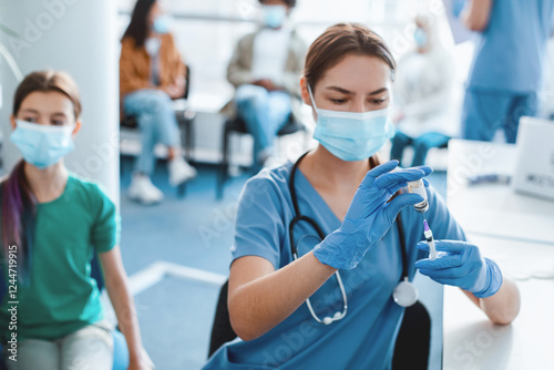 Coronavirus Immunization. Doctor With Syringe Taking Dose Of flu Vaccine For Injection From Vial, Holding Bottle Wearing Blue Latex Gloves And Protective Mask, Kid Waiting In Background