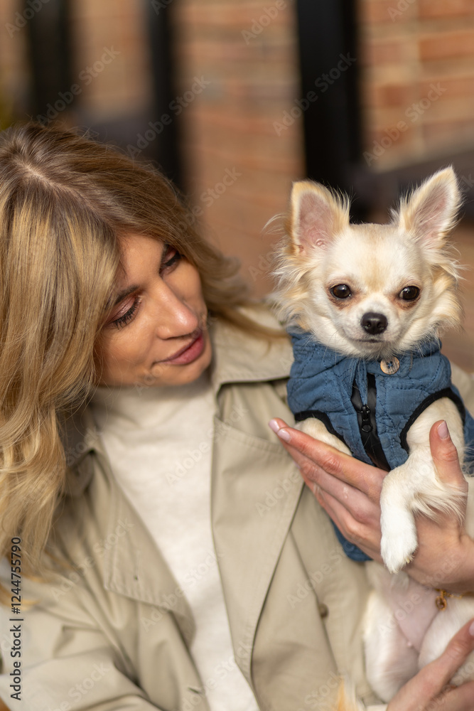 Woman holds small dog in denim jacket while smiling warmly at the camera in a cozy outdoor setting Woman holds small dog in denim jacket while smiling warmly at the camera in a cozy outdoor setting