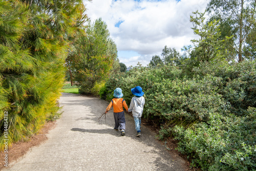 Fototapeta Naklejka Na Ścianę i Meble -  Two young children (3 to 4 years old) wearing wide brim sun hats walk hand in hand along a public park pathway in Australia. A heartwarming moment of outdoor exploration and childhood friendship.