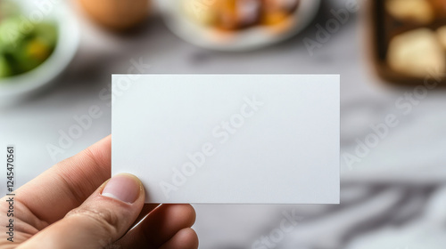 A close-up photograph of an empty white business card, held in a hand against a blurred background with some food items in the background